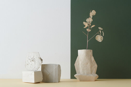 Horizontal shot of creative color-blocking still life composition of gypsum vessel, while objects and dried plant against white and dark green wall background, copy space