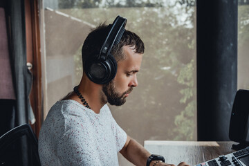 young handsome man with a beard and dark hair sits at a table and creates music, listens to headphones, writes songs, musician portrait, concept, laptop, monitor speakers, pianist, recording studio