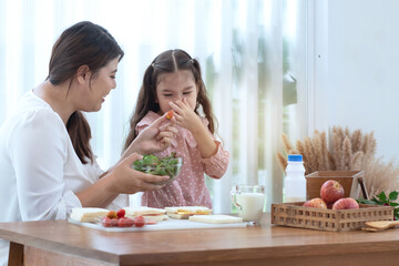Little girl shows a gesture of covering her nose with her fingers while her mother sent vegetables to try, don't want to eat vegetables - obrazy, fototapety, plakaty