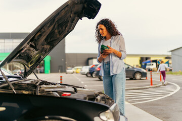 Obraz premium A young woman stands in front of a car with the hood up and reads the instructions on how to change the oil