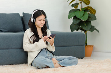 Asian young happy cheerful female model wearing stereo headset headphone sitting smiling on fluffy carpet floor using touchscreen smartphone listening to music playlist song in living room at home
