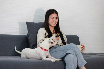 Asian young happy cheerful female owner sitting smiling on cozy sofa couch watching movie with best friend companion dog white short hair jack russell terrier