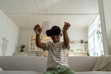 Little african american boy wearing 3d virtual reality glasses sits on couch at home. Online world...