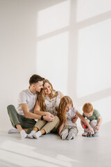 Family moving into new house. Relocation concept. Young happy mother, father, daughter, son isolated on white wall. Funny parents hug children sitting on floor at home. Childs play toys, girl and boy.