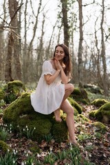 Portrait of a woman in the forest. She is sitting in a white dress on a meadow with snowdrops in a spring forest