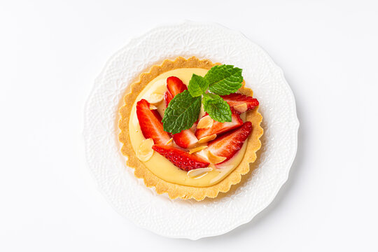 Overhead shot of strawberry mini cake tartlet with vanilla custard and mint leaves over white background. Concept of homemade sweet pastries. Copy space, top view