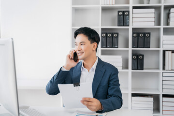 Asian businessman involved in cellphone call conversation working on computer, giving professional consultation to client or negotiating project.