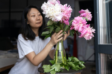 Asian woman puts peonies flowers in vase. Housewife enjoying decor and interior of kitchen. Sweet home. Allergy free.
