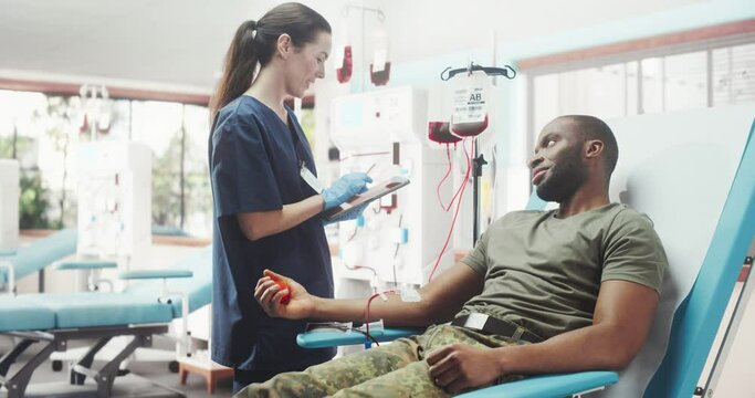 Caucasian Female Nurse Taking Blood Donation From Black Army Soldier In Military Hospital. African American Man Squeezing Heart-Shaped Ball To Pump Blood Into Bag. Troop Donating To Wounded Comrades.