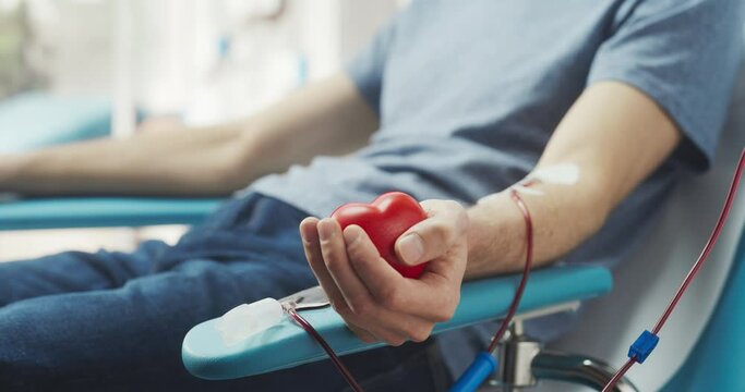 Close Up Shot Of Hand Of Male Blood Donor With an Attached Catheter. Caucasian Man Squeezing Heart-Shaped Red Ball To Pump Blood Through The Tubing Into Bag. Donation For Victims Of Accidents.