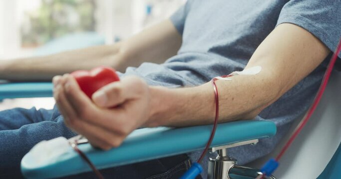 Close Up Shot Of Hand Of Male Blood Donor With Attached Catheter. Male Squeezing Heart-Shaped Red Ball To Pump Blood Through The Tubing Into Bag. Donation For Patients Requiring Blood Transfusions.