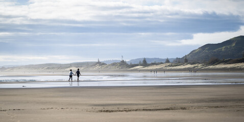 Fototapeta na wymiar Couple holding hands and walking barefoot on Muriwai beach. Unrecognizable people walking in the distance. Auckland.