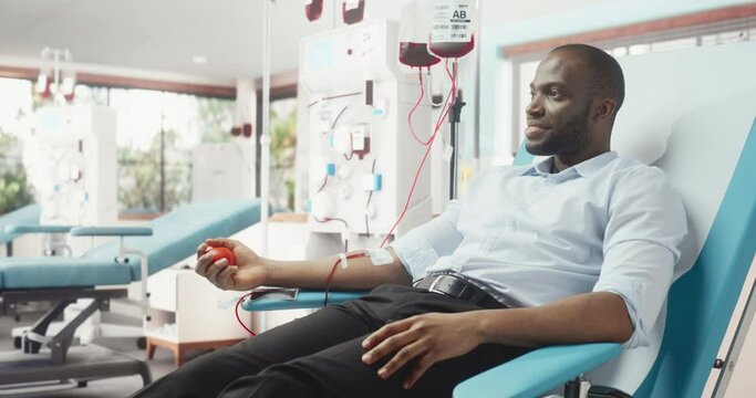 Black Entrepreneur Donating Blood For People In Need In Bright Hospital. Male Donor Squeezing Heart Shaped Red Ball To Pump Blood Through The Tubing Into The Bag. Donation For Burn Victims Patients.
