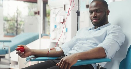 Black Businessman Donating Blood For People In Need In Hospital. African Male Donor Squeezing Heart-Shaped Ball To Pump Blood. Looking Into Camera And Smiling. Donation For Organ Transplant Patients.