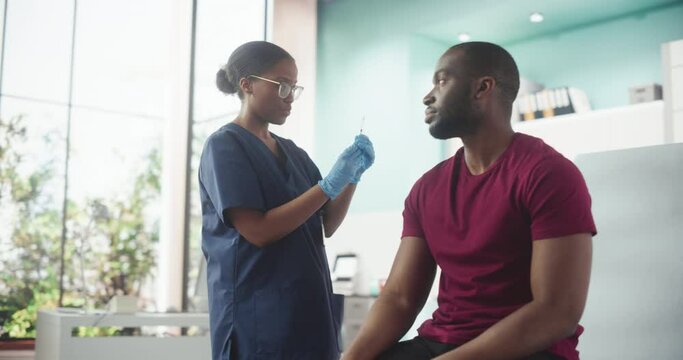 African American Man Sitting In The Chair In Hospital And Getting His Chicken Pox Vaccine. Professional Black Female Nurse Is Performing Injection And Putting A Patch On. Public Healthcare Concept.