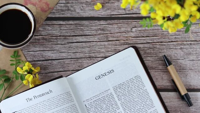 Hands flipping pages of open holy bible book Genesis with cup of coffee, pen, and sping flowers on wooden table. Top view. Reading and studying Scriptures, Christian education, wisdom from God.