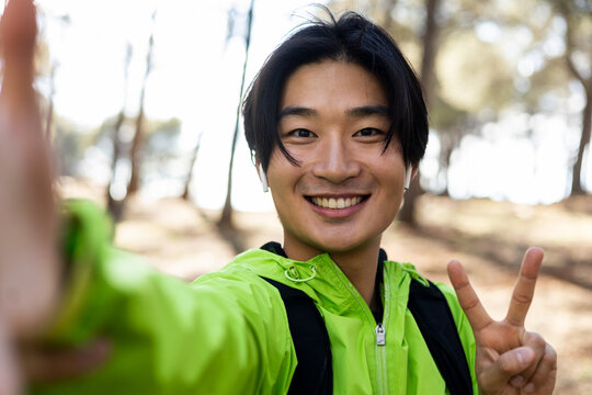 A cute Asian is taking a picture with his smart phone at the top of the mountain. The smiling young man makes the victory gesture with his fingers. Hiking concept, Asian excursion. Spring holidays.