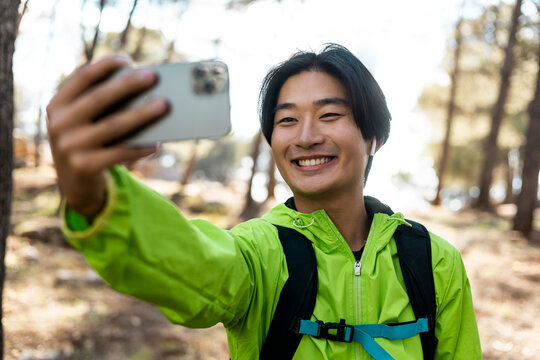 A cute Asian is taking a picture with his smart phone at the top of the mountain. The young man uploads photos for social networks. Hiking concept, smiling Asians excursion. Spring holidays.