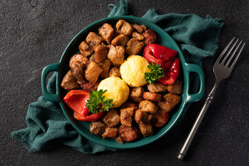 Romanian food pomana porcului made from chunks of fried pork and sausages served with corn porridge and pickled red peppers in a plate on a dark background. Horizontal Top View.