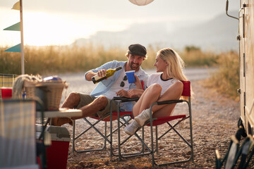 Couple enjoying summertime camping with camper rv in nature, sitting and pouring a glass of wine. Fun, travel, togetherness, lifestyle, love concept.