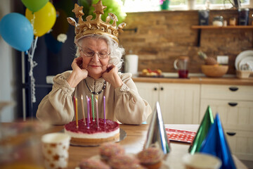 Elderly elegant gray haired senior woman wearing a golden crown celebrating her birthday, sitting in front of birthday cake.