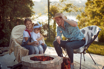 Joyful young girls roasting marshmallows by a fireplace with their dad outdoors in a cozy cottage, enjoying sunny summer day together.
