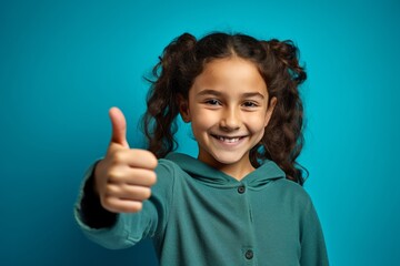 Close-up portrait photography of a grinning kid female showing a thumb up against a turquoise blue background. With generative AI technology