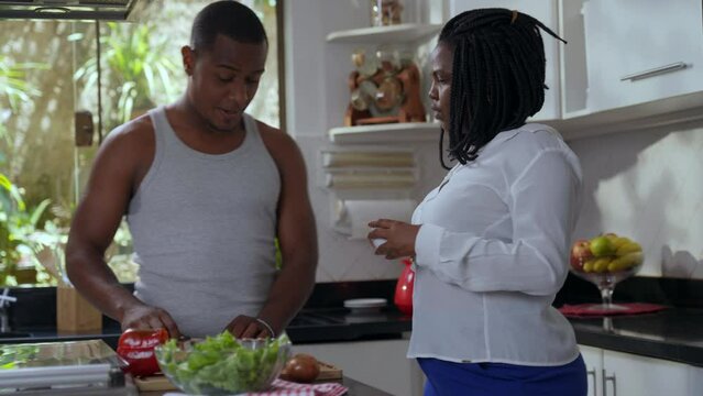 brazilian black woman drinking coffee in the kitchen and talking to hier boyfriend while he cuts vegetables for his valentine's day lunch