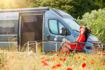 Work and Travel concept, outdoor leisure activity and wanderlust lifestyle - woman dressed in red dress resting in a chair in a meadow among blooming red poppies next to an off-road camper van.