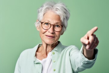 Close-up portrait photography of a glad mature woman making a i have an idea gesture with a finger up against a pastel green background. With generative AI technology