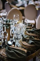 Round served banquet tables for guests, decorated plate, glasses, forks, emerald green napkins. Elegant dinner table. Wedding hall with atmospheric decor. Golden geometric decorative elements decor.
