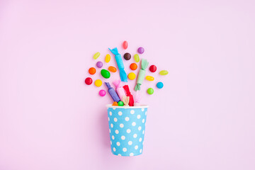 Different kinds of colorful candy out of a blue paper cup with white dot on pink background,...