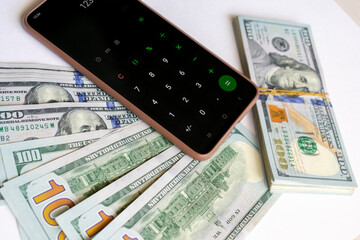 Fototapeta premium A stack of one hundred dollar bills and a calculator on a smartphone. A stack of one hundred dollar bills and a smartphone on a light background.