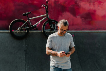 Portrait of a middle-aged contemporary tattooed man standing in a skate park and typing on his...
