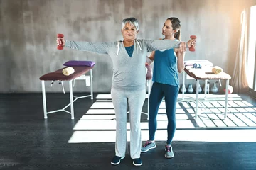 Poster Physical therapy, dumbbell and coaching with old woman and personal trainer for support, health and physiotherapy. Training, weightlifting and fitness with senior patient and female trainer for help © Mikolette Moller/peopleimages.com