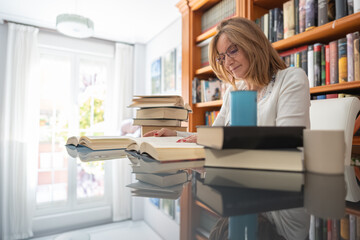 Mature white woman with several books on a glass table and daylight pouring in through the window.