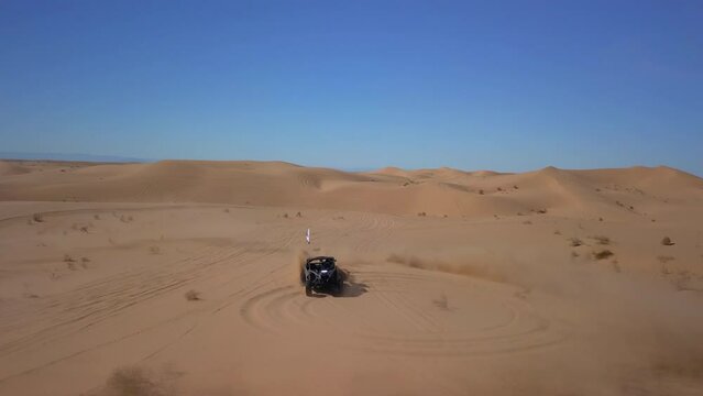 Aerial: Drone Flying Over Dune Buggy Doing Doughnut On Sand Amidst Bushes Against Blue Sky - Glamis, California