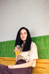 Portrait of a cute casual girl drinking orange juice from a glass and looking in cafe, cafeteria, restaurant on green tile background.