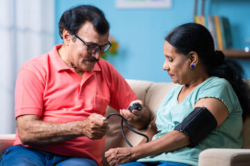 Indian senior man checking blood pressure or bp of wife by using sphygmomanometer while sitting on...