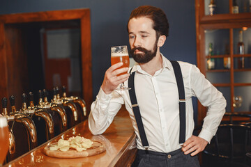 Man with a mustache and beard stands at the bar and drinks alcohol from a glass.