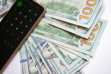 A stack of one hundred dollar bills and a calculator on a smartphone. A stack of one hundred dollar bills and a smartphone on a light background.