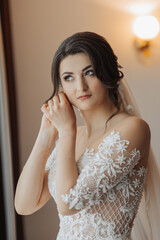 A brunette bride in a white voluminous dress with long lace sleeves, puts on her earrings, poses in her room. Great makeup. Open shoulders. Beautiful hands. Long veil. Morning of the bride. Portrait