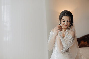 A brunette bride in a white voluminous dress with long lace sleeves, puts on her earrings, poses in her room. Great makeup. Open shoulders. Beautiful hands. Long veil. Morning of the bride. Portrait