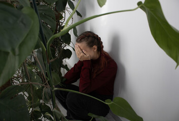 A teenage girl is sitting with her face in her hands in the corner by the wall next to the houseplants. She's upset and sad. The problem of loneliness, bullying, mental health of adolescents in the mo