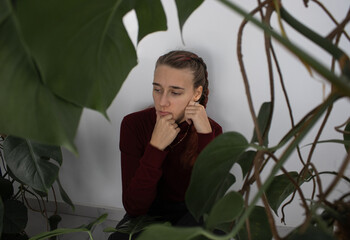 A girl is sitting in the corner by the wall in the room next to the houseplants. She's upset and sad. The problem of loneliness, bullying, mental health in the modern world.