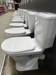 Samples of сeramic white toilet bowls standing in a row in the warehouse of a plumbing store. Samples of modern sanitary ware for the toilet. New modern toilet in the plumbing store