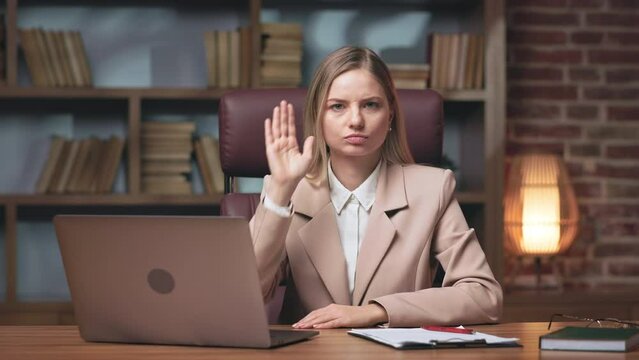 Front view of determined young businesswoman sitting alone at office table with laptop and showing stop gesture. Strict female holding arms crossed and saying no to coworkers.