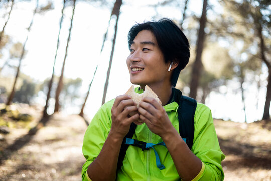 A young Asian is hiking up a mountain. The smiling young man is eating a sandwich while looking to the side. Nature concept, Asian excursion. Take a tentempie in the forest.