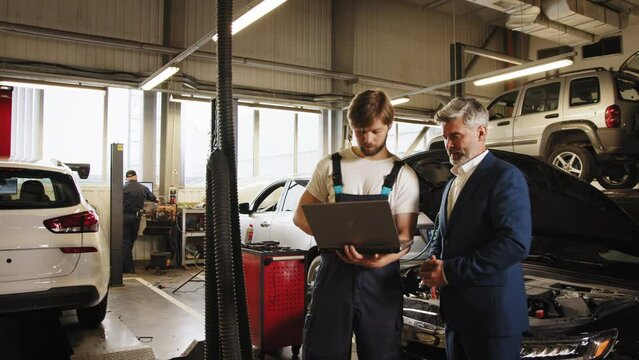 Auto mechanic showing to a male customer the engine error codes in auto-service. Handsome male mechanic wearing uniform, using laptop talking with grey hair business man client.