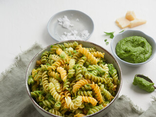 close up view of bowl with colored fusilli pasta and pesto sauce. In the background out of focus parmesan cheese and basil leaves.